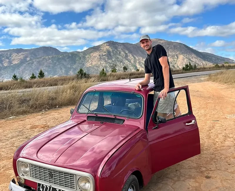 Een reiziger staat in de deur van een Renault 4 op een rode zandweg