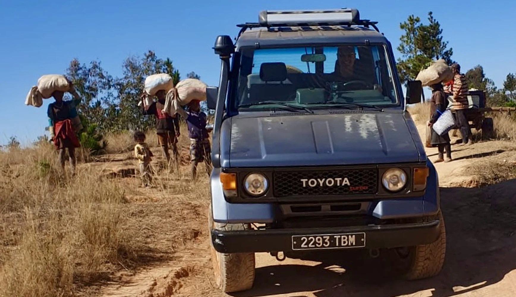 Notre Toyota Land Cruiser bleu sur une piste, tandis que des villageois portent des sacs sur la tête
