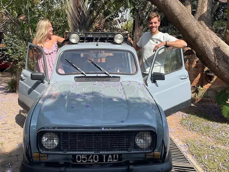 Two travellers stand in the open doors of a pale blue Renault 4 in a garden