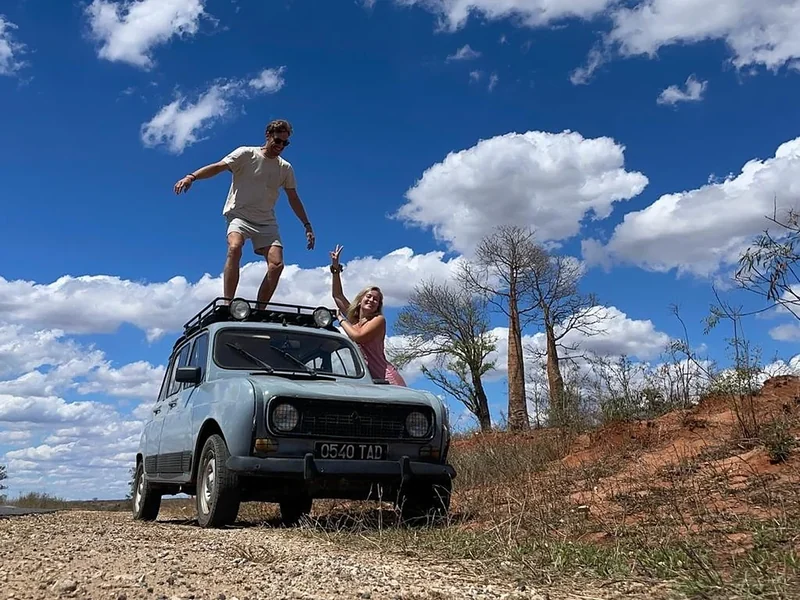 Een reiziger staat op het dak van een blauwe Renault 4 tussen de baobabs