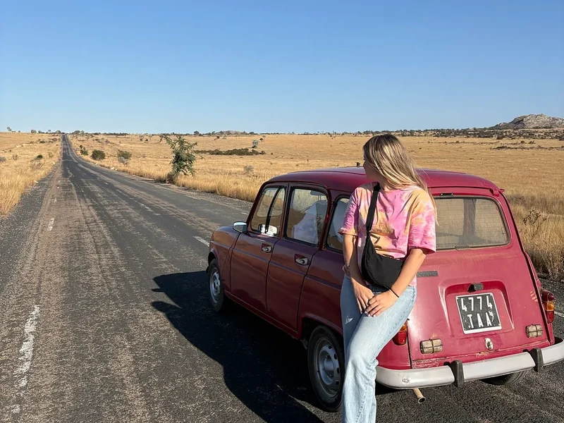 A traveller leans on a pink Renault 4 on an empty road