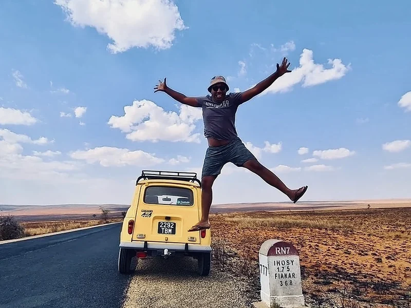 A traveller jumps by a yellow Renault 4 next to an RN7 kilometre stone
